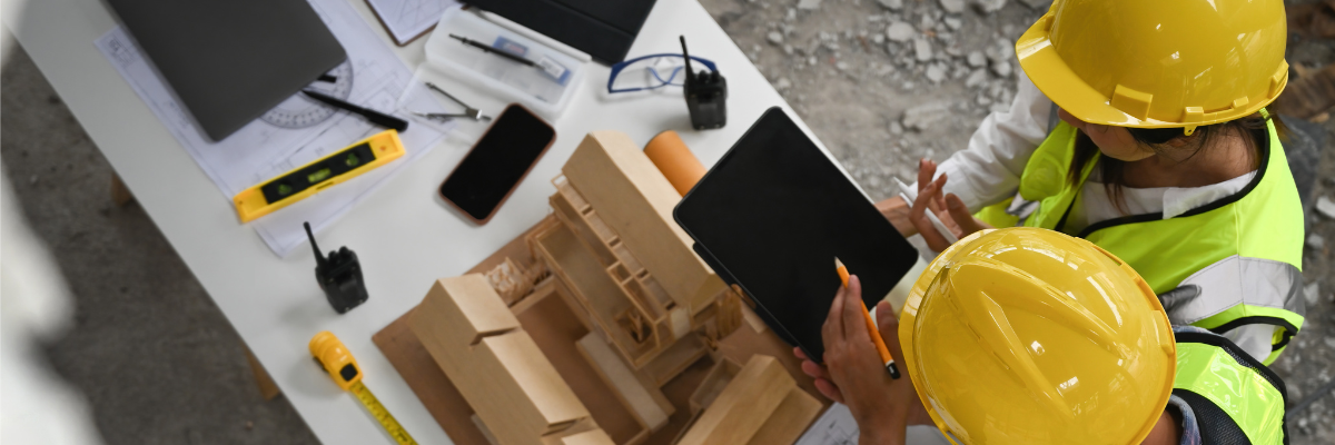 Two people in yellow construction helmets and safety vests review a tablet above a desk with architectural models, blueprints, measuring tools, and electronic devices on a construction site.